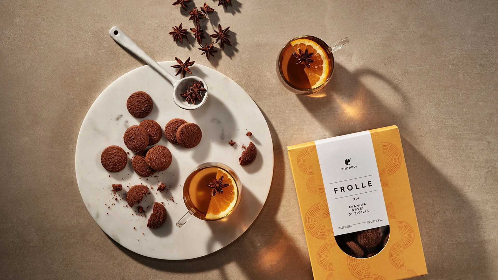 Chocolate cookies on a white marble plate with orange-tea glasses and a yellow cookie box nearby, styled on a brown surface with star anise garnish.
