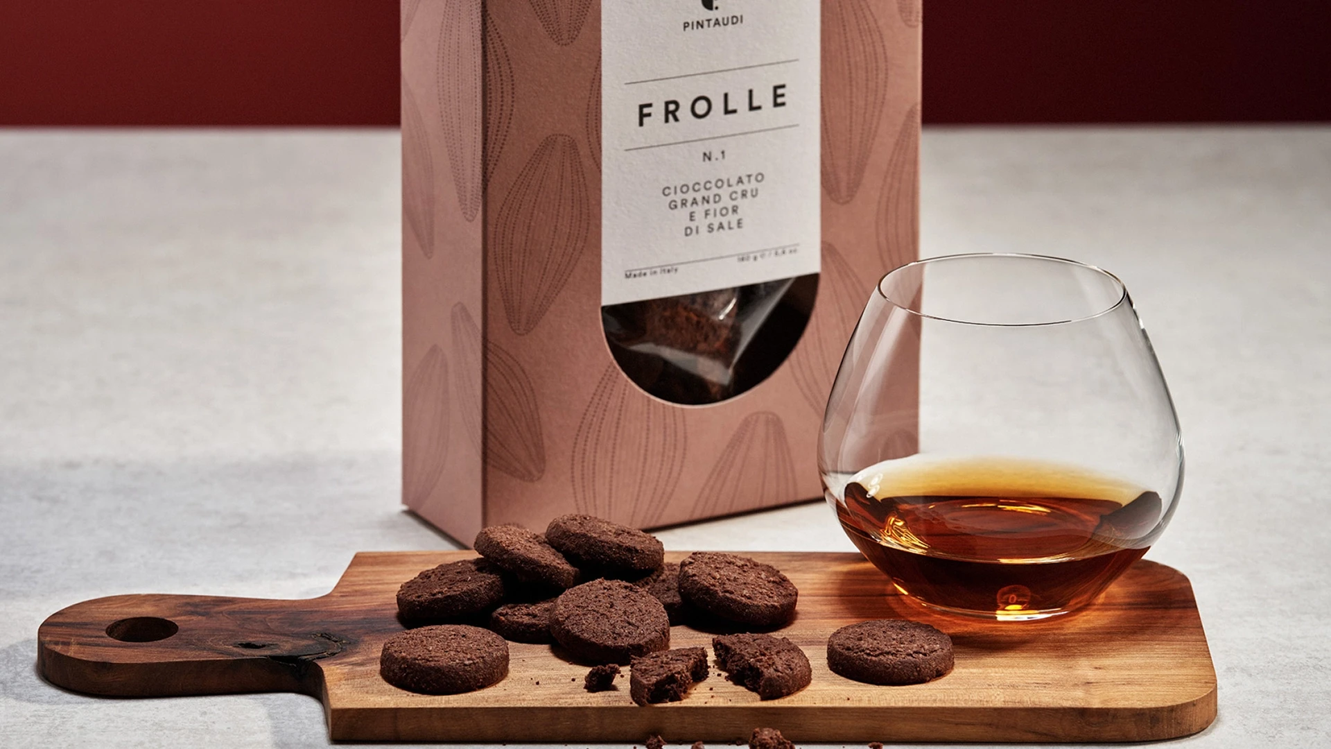 Chocolate cookies scattered on a wooden board next to a pink box labeled 'FROLLE' and a glass of amber whiskey or liqueur in the foreground.