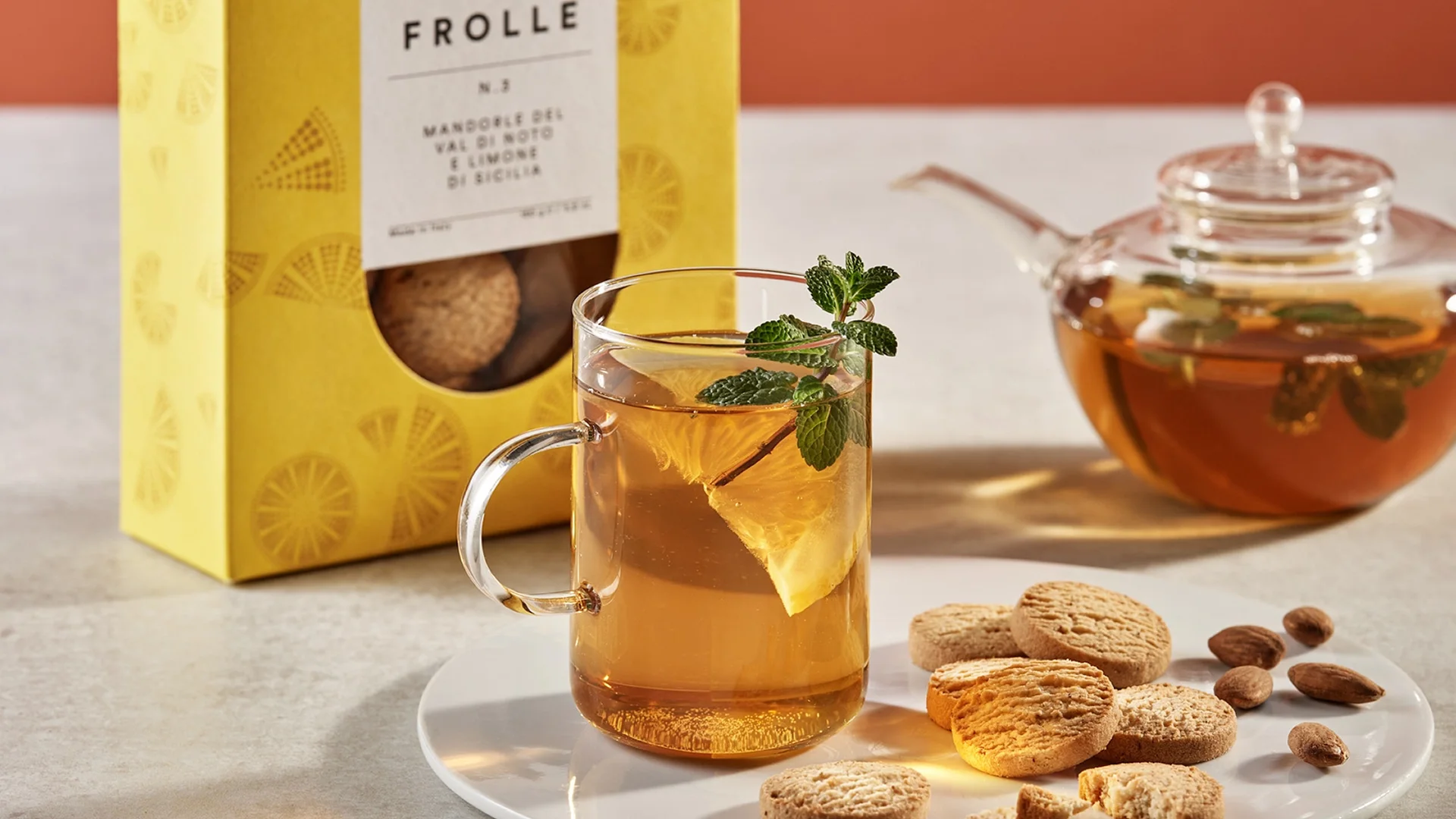 Glass mug of lemon-mint tea with a lemon wedge and fresh mint, on a plate with cookies nearby; a yellow packaging box in the background.