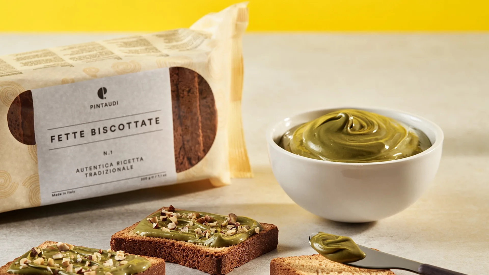 Package of PINTAUDI Fette Biscottate beside a bowl of pistachio spread and prepared toasts with chopped nuts on a light counter.