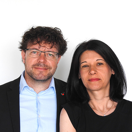 Man with glasses and beard in a blue shirt and dark blazer, standing next to a woman with black hair in a black top, against a white background.