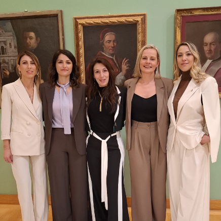 Five women in professional outfits stand side by side in front of framed portraits on a gallery wall.