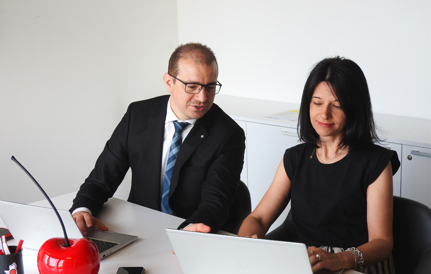 Two colleagues in a modern office collaborate at a white desk, each focused on their laptops.