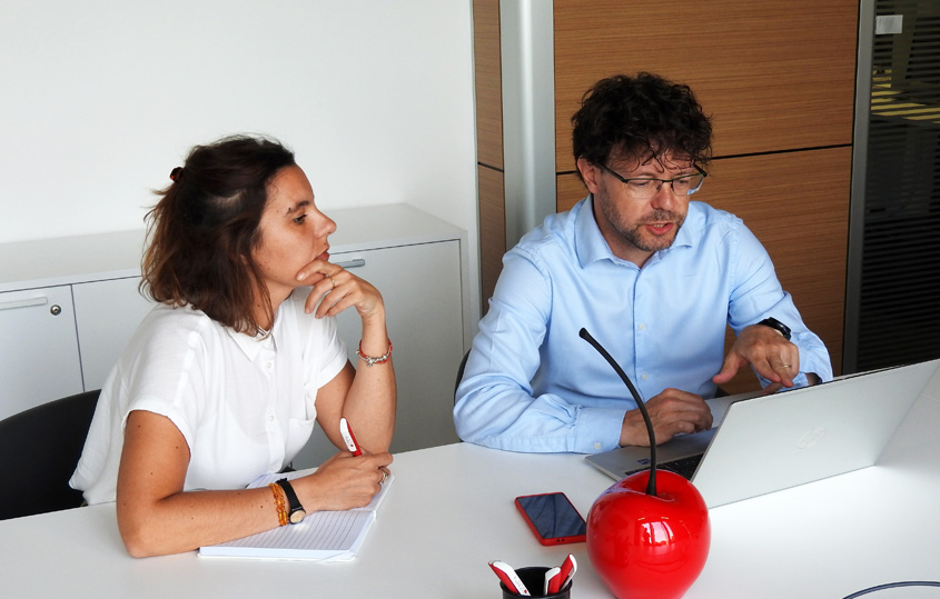 Two colleagues sit at a white table in a modern office; the man in a blue shirt uses a laptop while the woman takes notes.