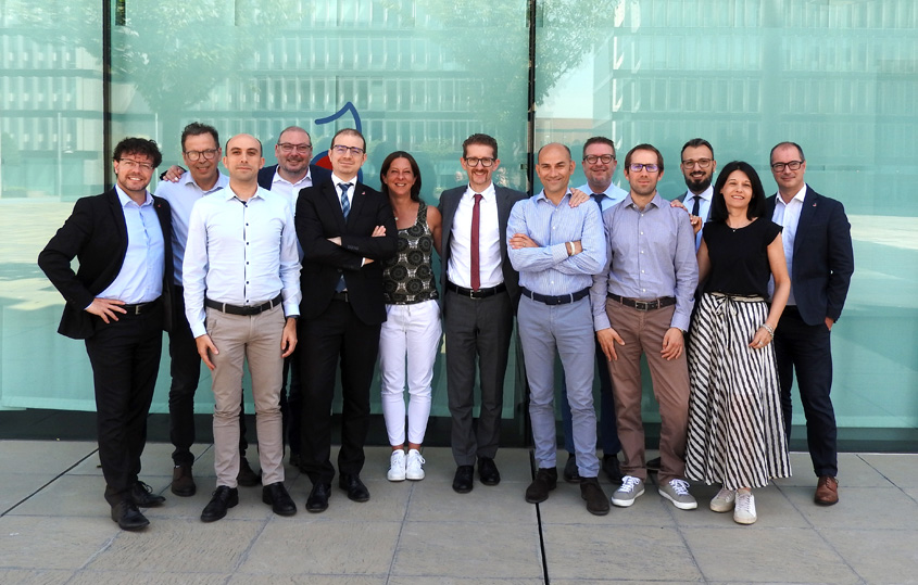 Group of eleven professionals standing together outside a glass office building, smiling at the camera.