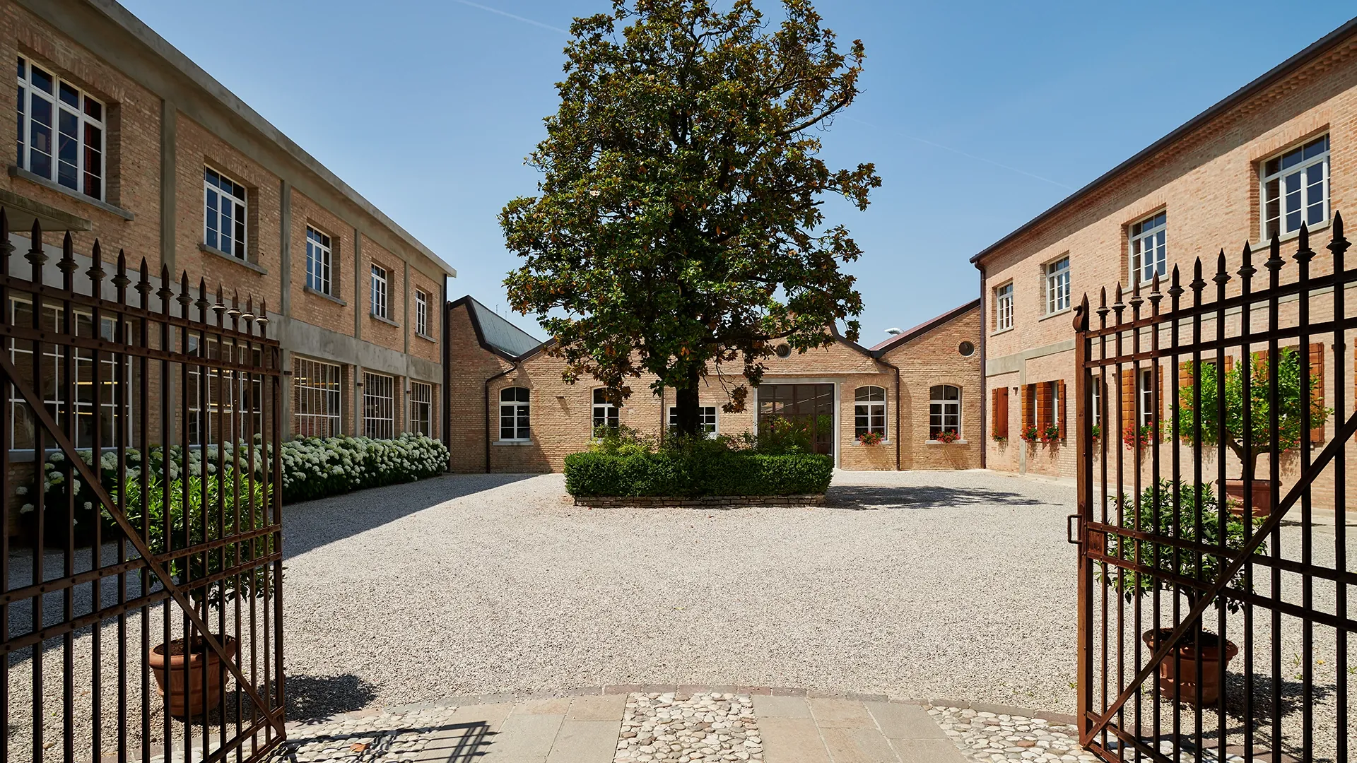 Open wrought-iron gate leads into a brick courtyard with a large central tree and two symmetrical brick buildings surrounding it.