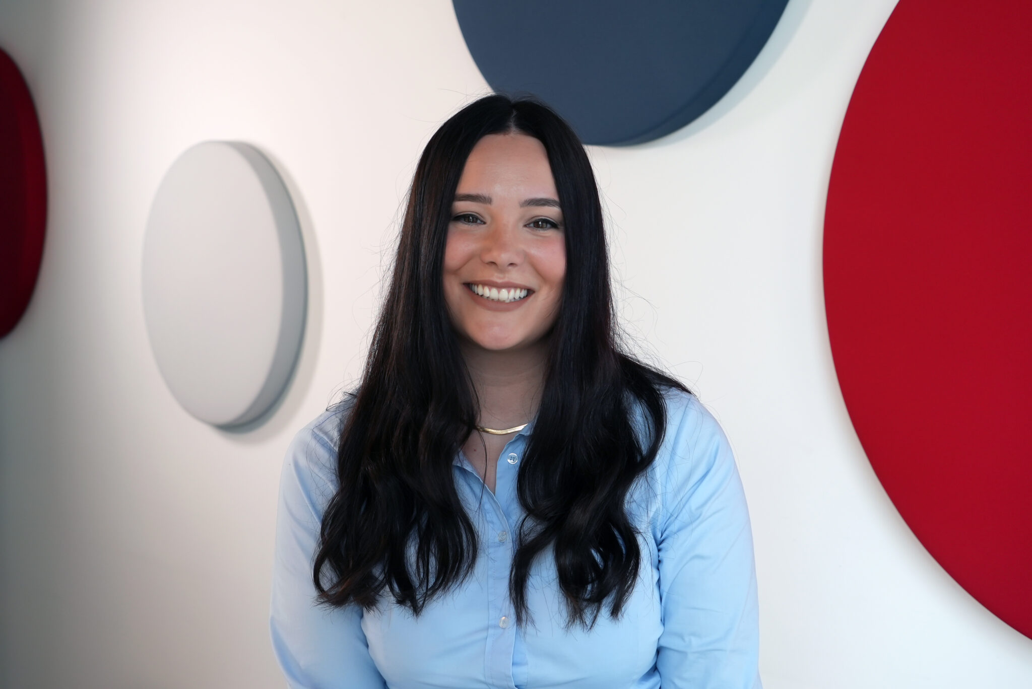 Smiling woman with long black hair in a light blue shirt, standing before white walls with circular wall decor (blue and red).