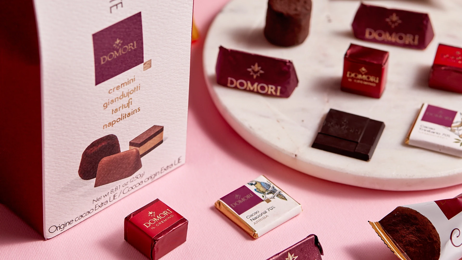 Assortment of Domori chocolates on a white marble tray with a Domori box in the foreground, set on a pink background.