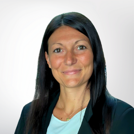 Portrait of a woman with long dark hair, wearing a blazer and light blue top, smiling against a pale studio backdrop.