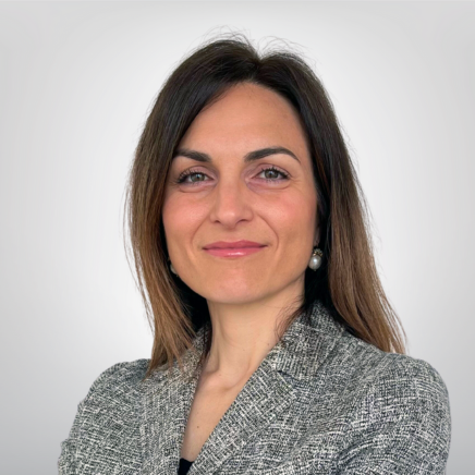 Professional headshot of a smiling woman in a gray blazer against a light neutral background.