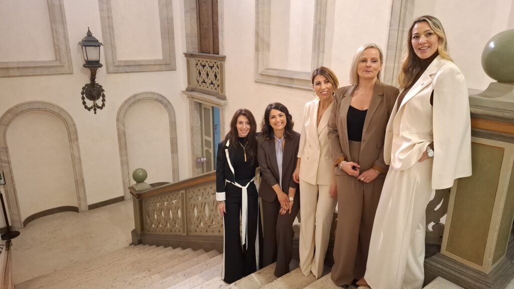 Five women in neutral-toned business outfits posing on a marble staircase inside a grand historic building.
