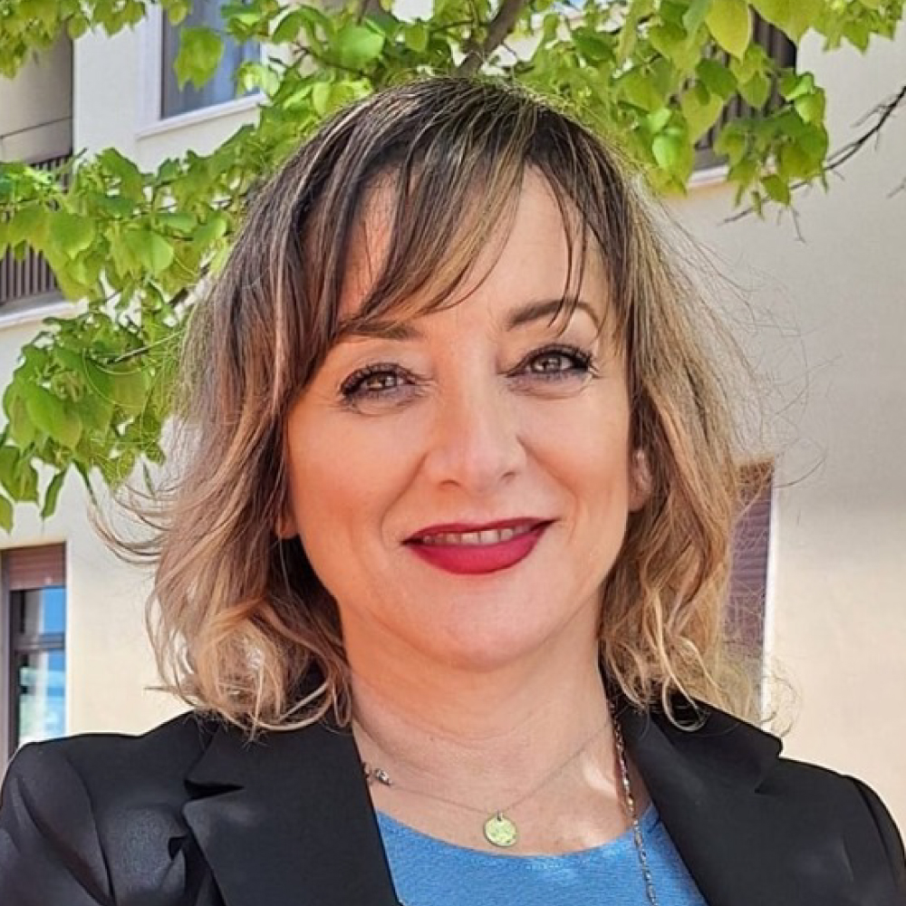 Smiling woman with shoulder-length light brown hair in a blue top and black blazer, outdoors with green leaves in the background.