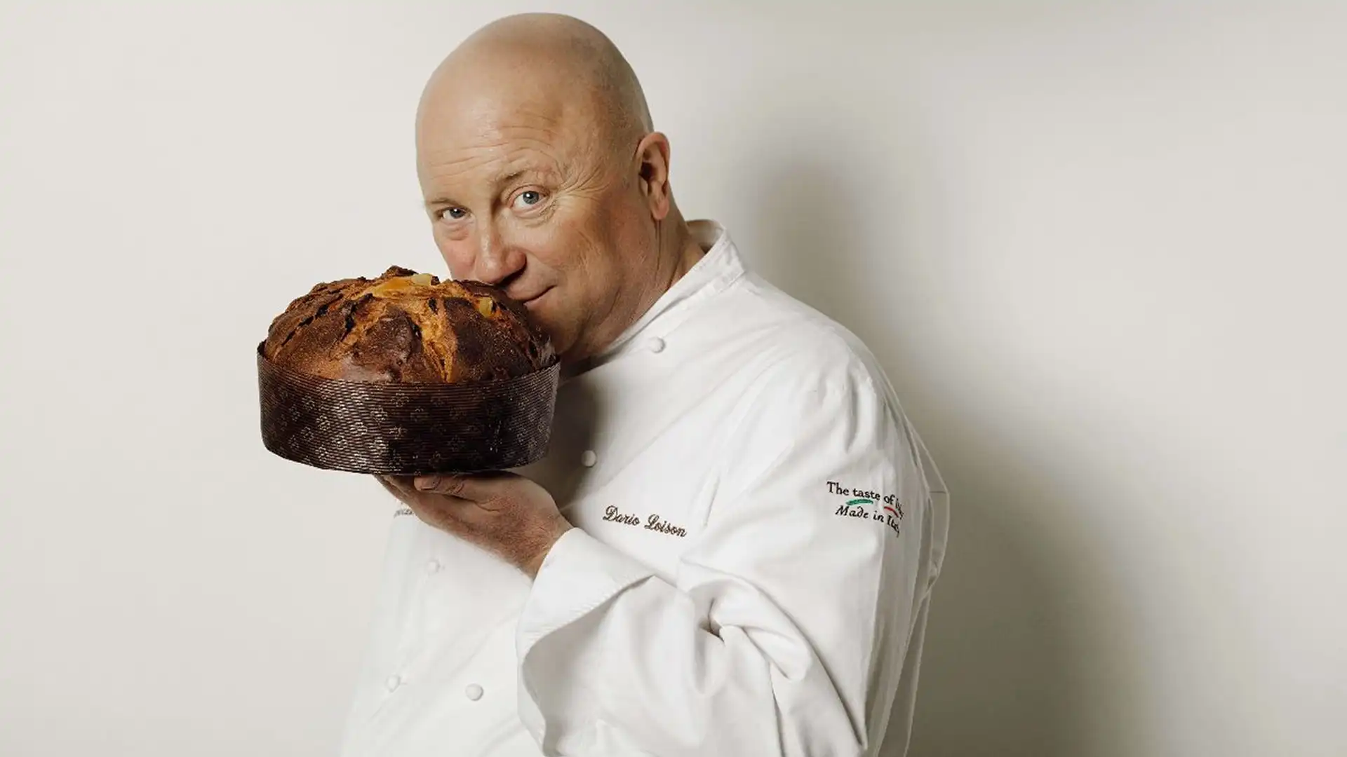 Chef in a white coat holding a round panettone up to his face, smiling at the camera in a studio setting