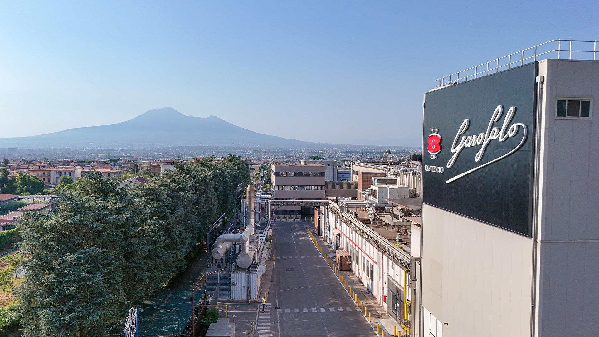 Industrial building with a large Garofalo logo on the side; pipes and a road in the foreground, city and distant hills beyond.