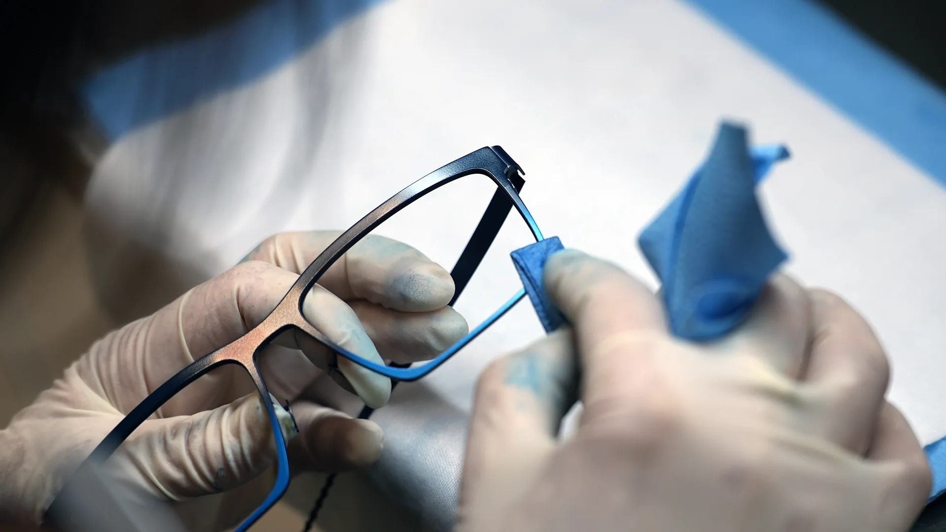 Close-up of gloved hands repairing a blue eyeglass frame with a cleaning cloth
