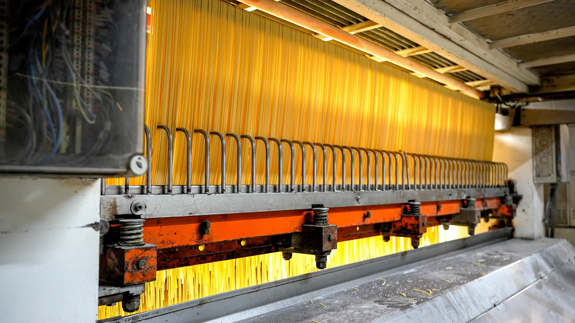 Industrial machine weaving yellow fibers, metal hooks align the strands above a red-orange frame in a factory setting