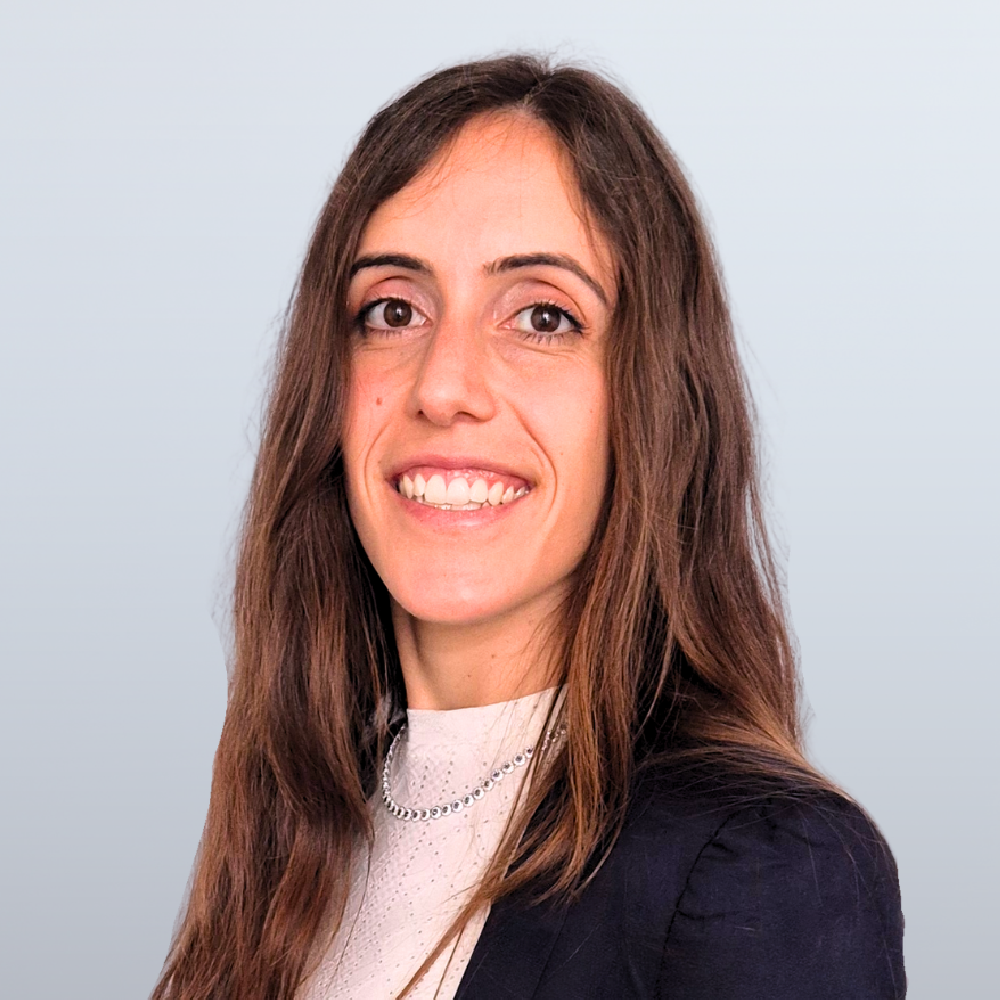 Smiling woman with long brown hair in a dark blazer and white blouse, portrait against a light blue background.