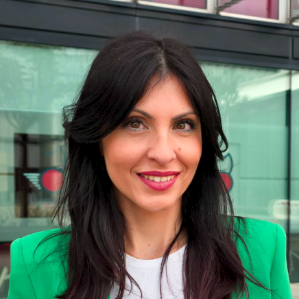 Portrait of a smiling woman with long dark hair in a bright green blazer, standing in front of a glass storefront.