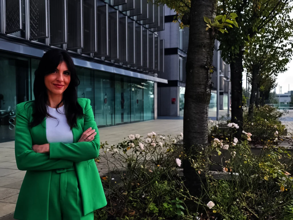 Woman in a bright green suit stands with arms crossed outside a modern glass-front building, smiling at the camera.