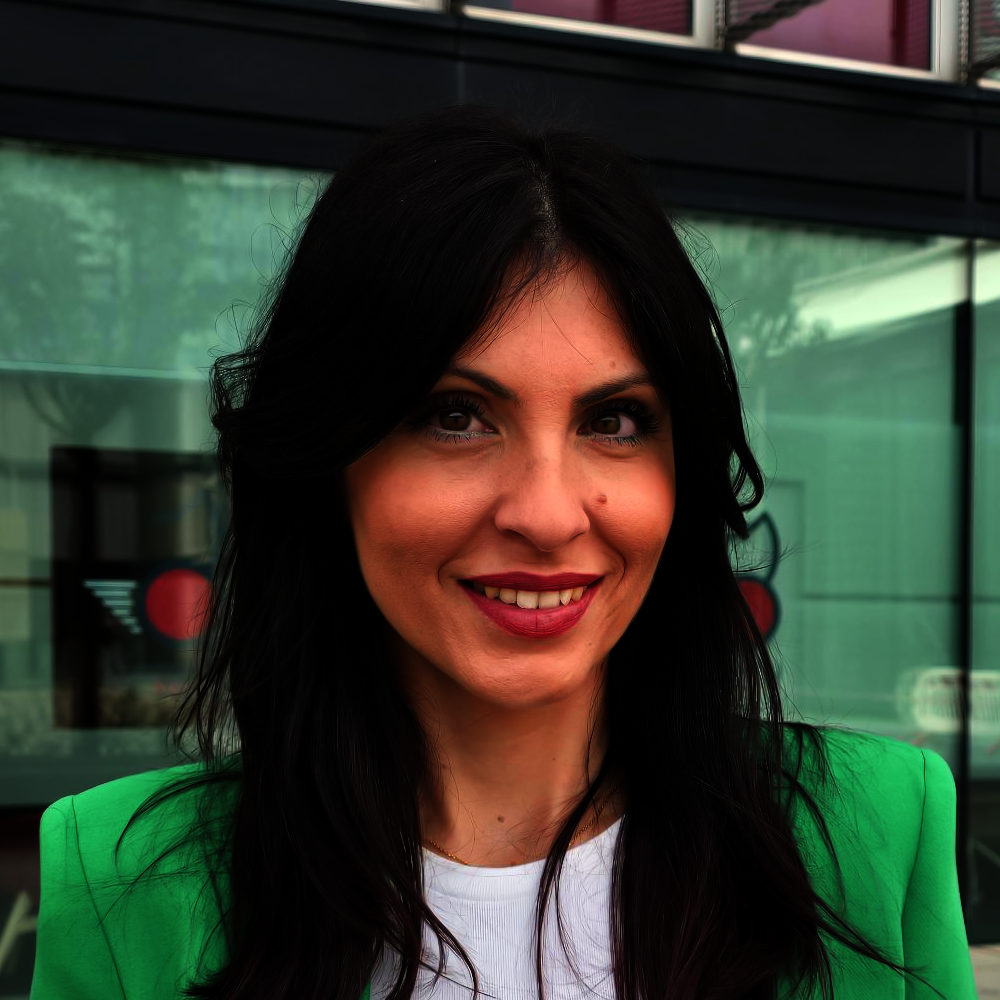 Portrait of a smiling woman with long dark hair, wearing a bright green blazer, standing outdoors in front of a glass building.