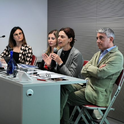 Panel discussion with four professionals seated at a table; the woman in a gray blazer speaks into a microphone while others listen.