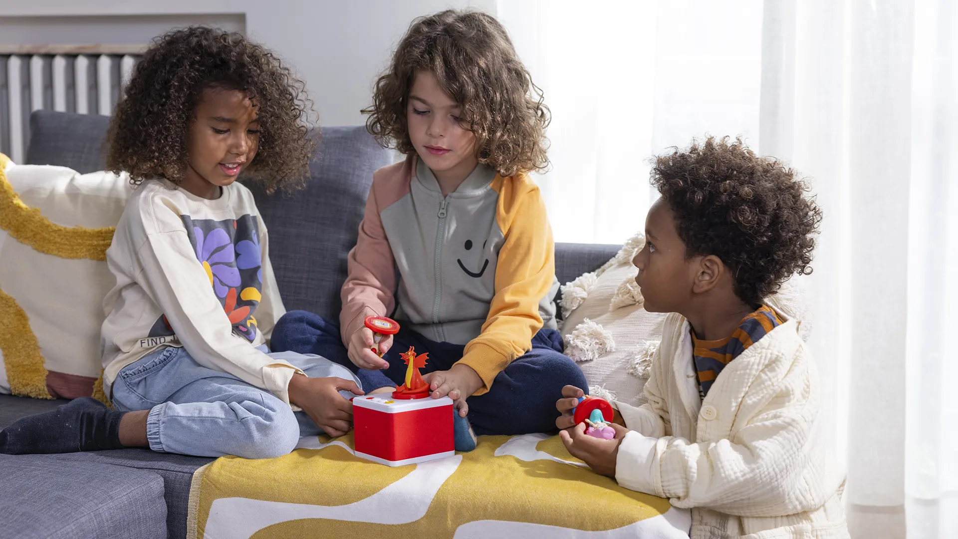 Three kids sit on a gray sofa in a sunlit room, playing with a red toy box and small figures on a yellow blanket.