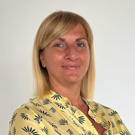 Portrait of a smiling woman with shoulder-length blonde hair wearing a yellow palm-patterned blouse, against a white background.