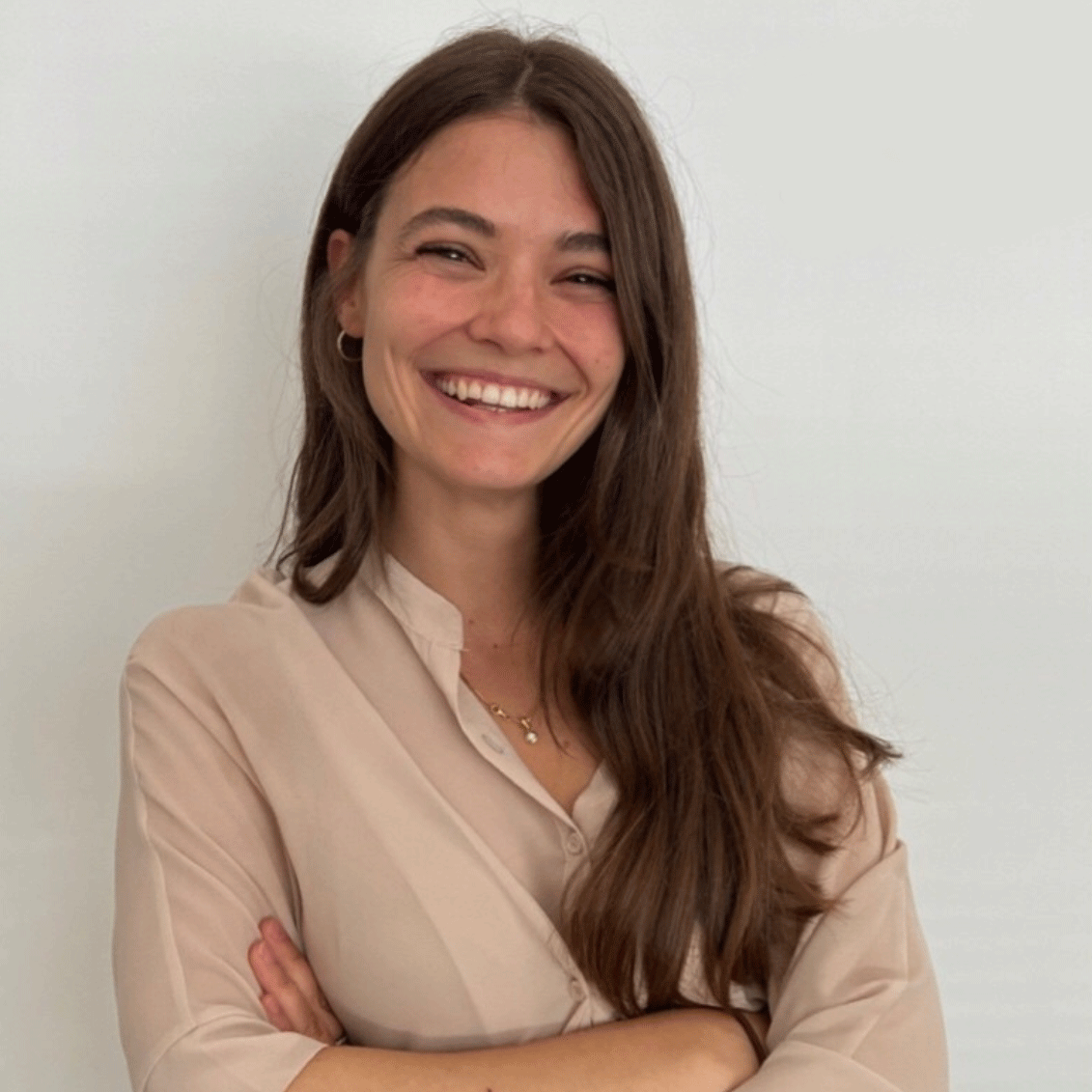 Smiling woman with long brown hair in a beige blouse, arms crossed, standing against a light wall.
