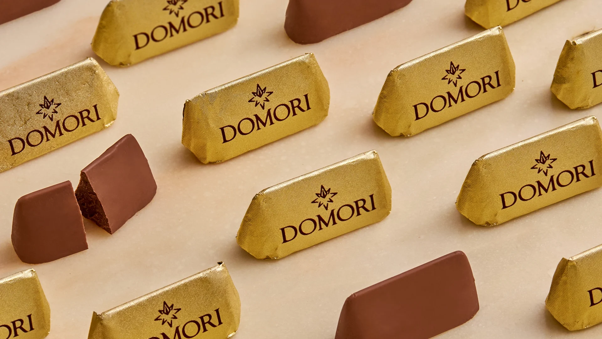 Gold-wrapped Domori chocolates scattered on a pale surface, with a few unwrapped pieces showing dark chocolate.