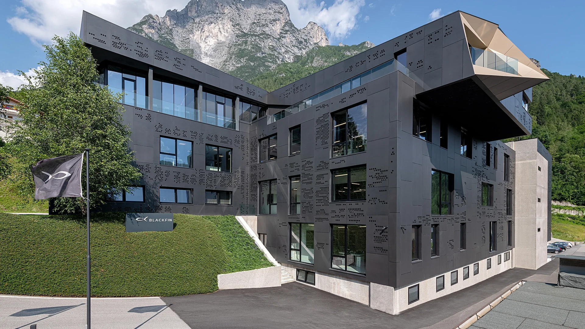 Modern dark-gray office building with perforated panels, large windows, and a mountain backdrop.