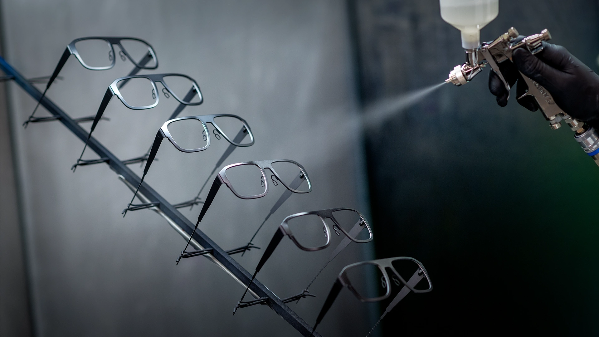 Eyeglass frames on a rack being spray-coated by a gloved technician in a workshop windowed area.
