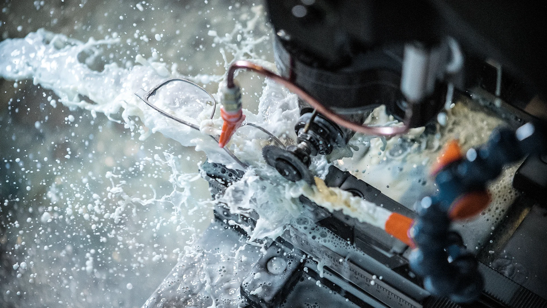 Close-up of a machining tool with coolant spray splashing around, glasses resting on the workpiece, and orange nozzles visible amid the motion.