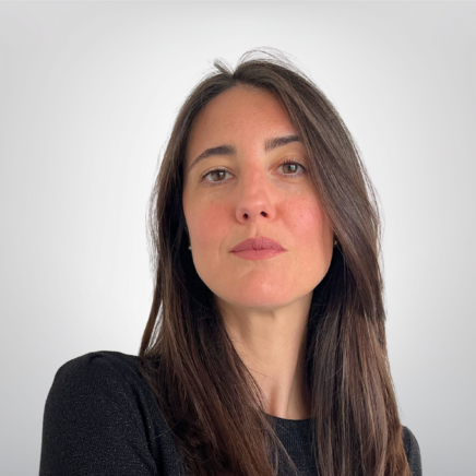 Portrait of a woman with long brown hair and a dark top, looking at the camera against a light background.