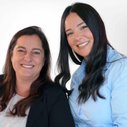 Two women posing together and smiling at the camera against a light background.