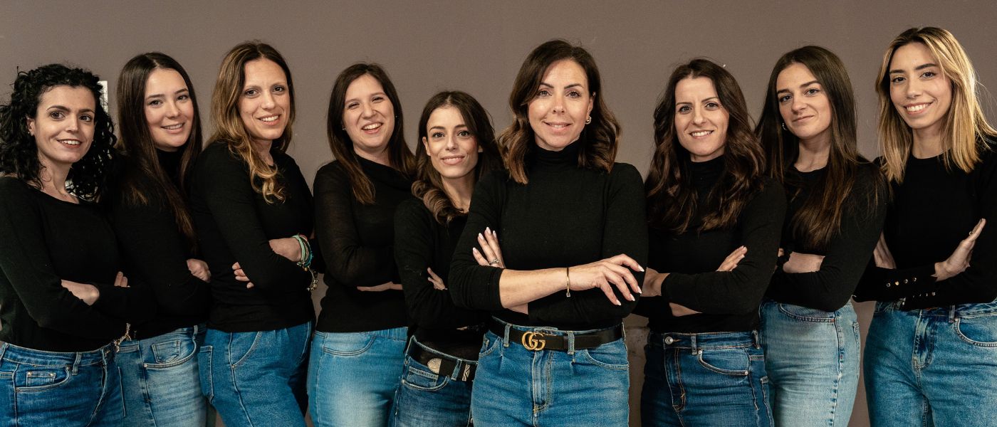 Group of nine women standing side by side in black tops and jeans, arms crossed, smiling at the camera against a neutral background.