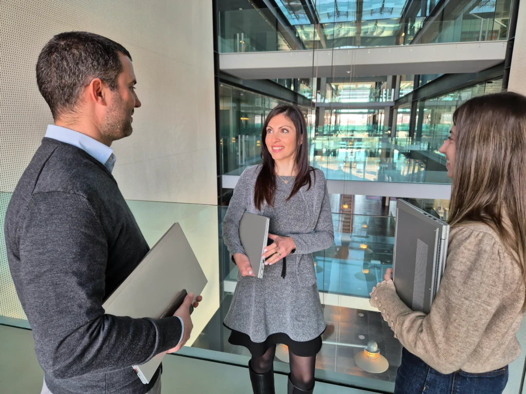 Three coworkers in a glass-walled atrium, each holding a laptop while talking.