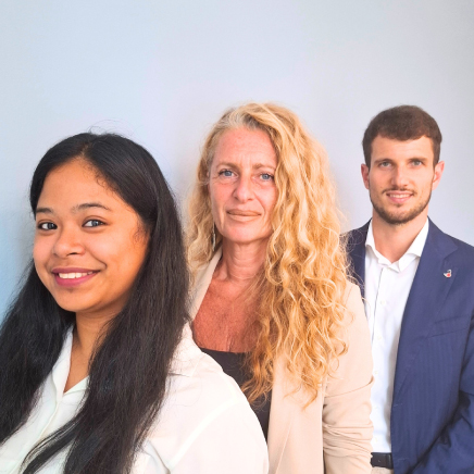 Group portrait of three professionals posing together: a woman with long dark hair front left, a blonde woman center, and a man in a blue blazer back right.