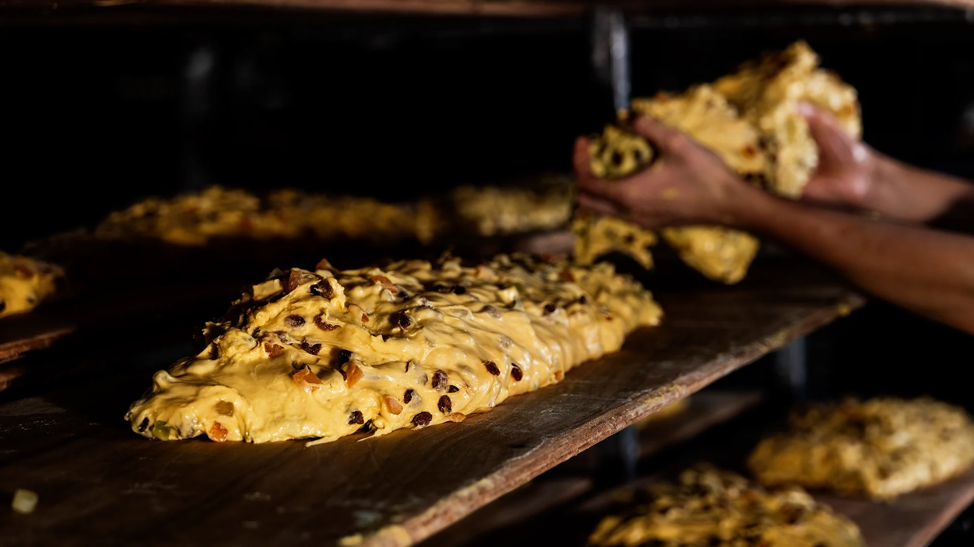 Raisin-filled cookie dough logs being shaped on a wooden baking board, hands working in the background.