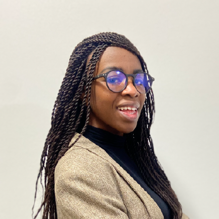 Portrait of a smiling Black woman with long braided hair, wearing round glasses and a beige blazer over a black top, against a light wall.