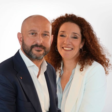 Two professionals posing for a business portrait: man in a navy suit and woman in a white blazer smiling.