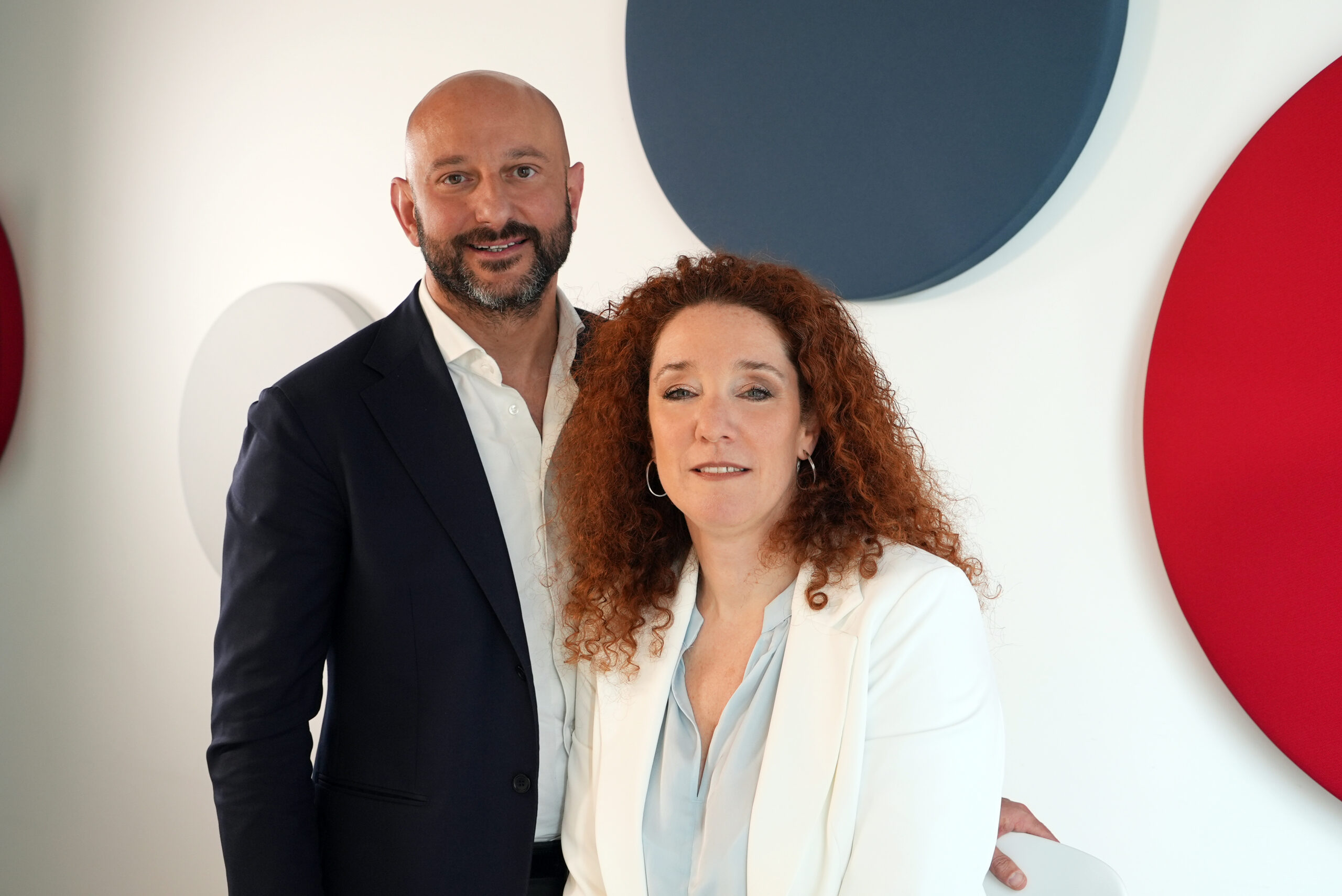 Two colleagues posing together indoors, man in a dark suit and white shirt beside a woman with curly red hair in a light blazer, abstract wall art behind them.