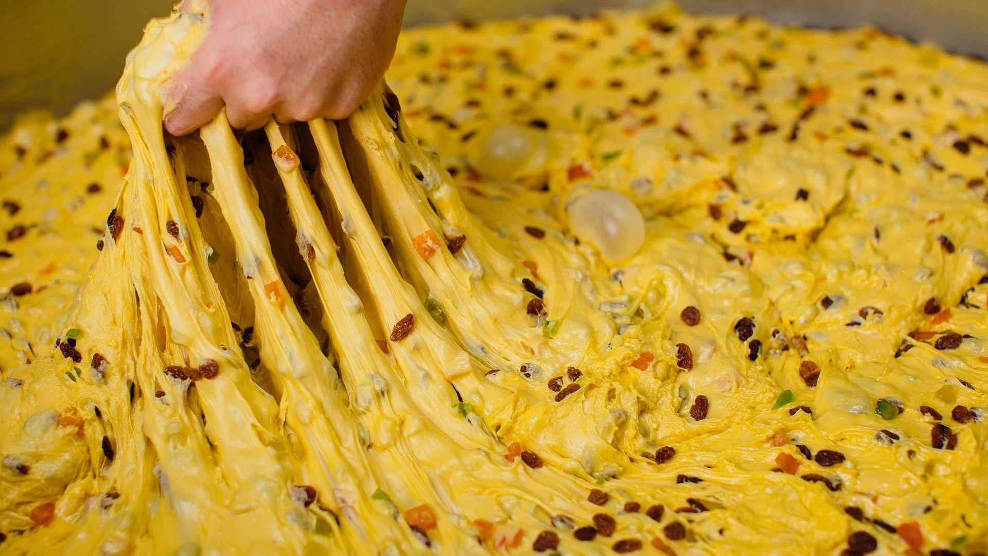 Close-up of a hand pulling apart a cheesy, pull-apart bread topped with colorful bits of vegetables and raisins.