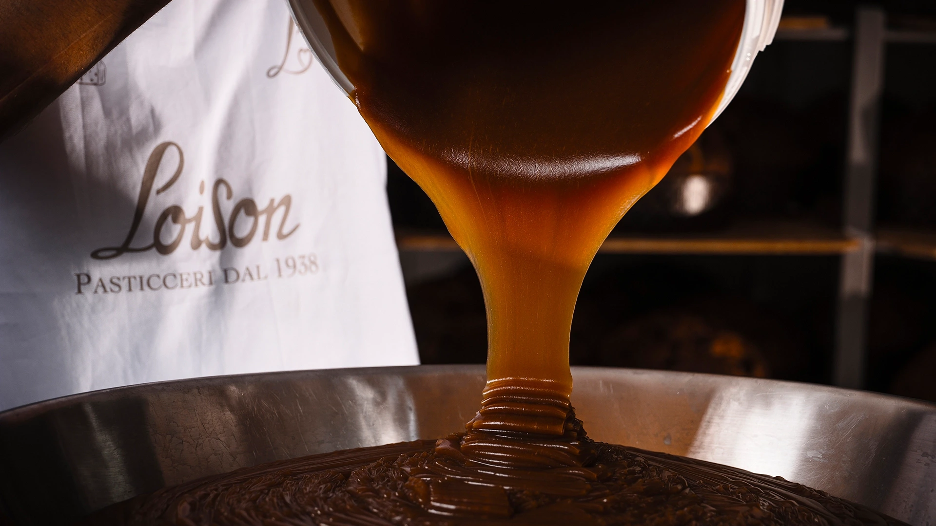 Close-up of dark chocolate being poured from a mold into a pan, with a baker's white apron in the background reading 'Pasticceri Dal 1938'.
