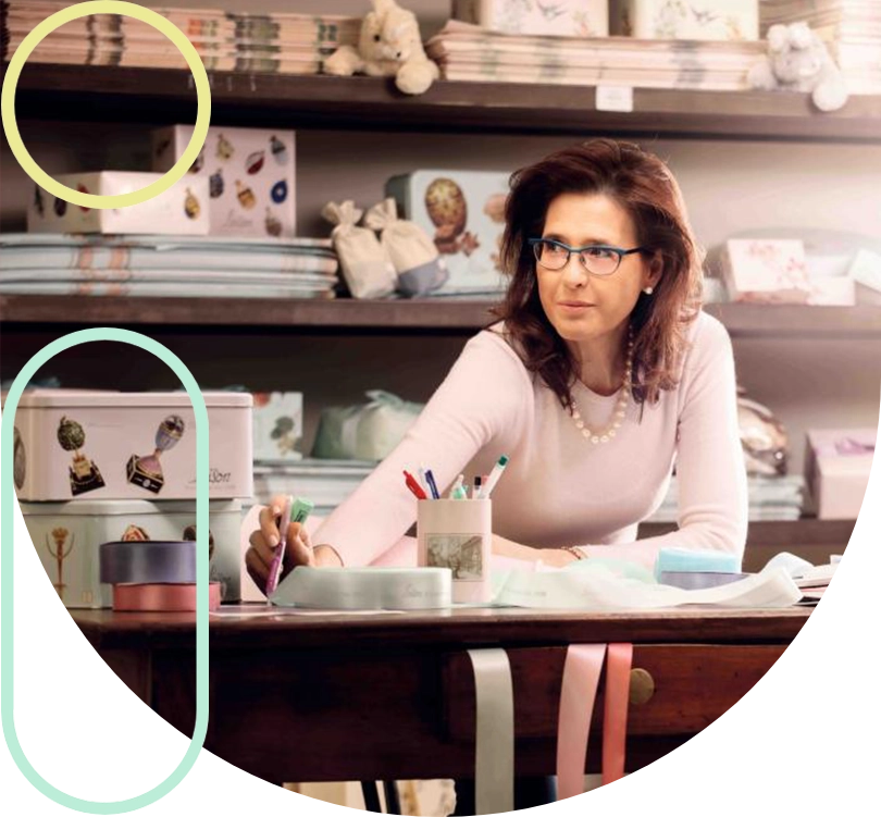 Store employee sits at a wooden craft desk, writing with a marker while shelves of gifts and supplies loom in the background.