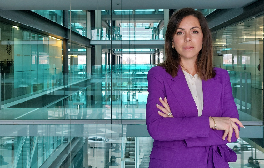 Businesswoman in a purple blazer stands with arms crossed in a glass-walled atrium.