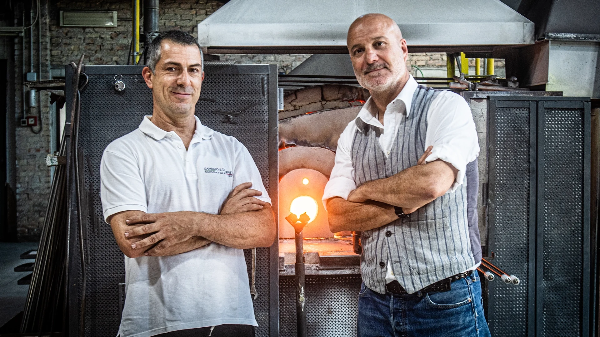 Two men with folded arms pose in front of a glowing furnace in a glassblowing workshop.
