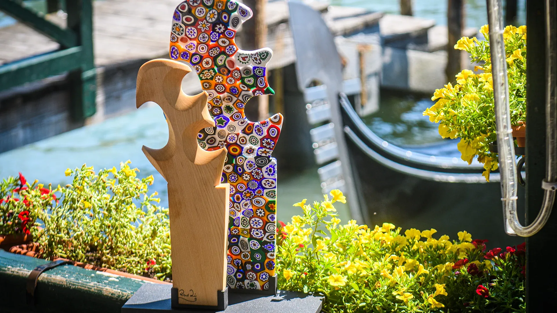 Two decorative sculptures on a pedestal among yellow flowers by a canal; one wood, one multicolored mosaic image.
