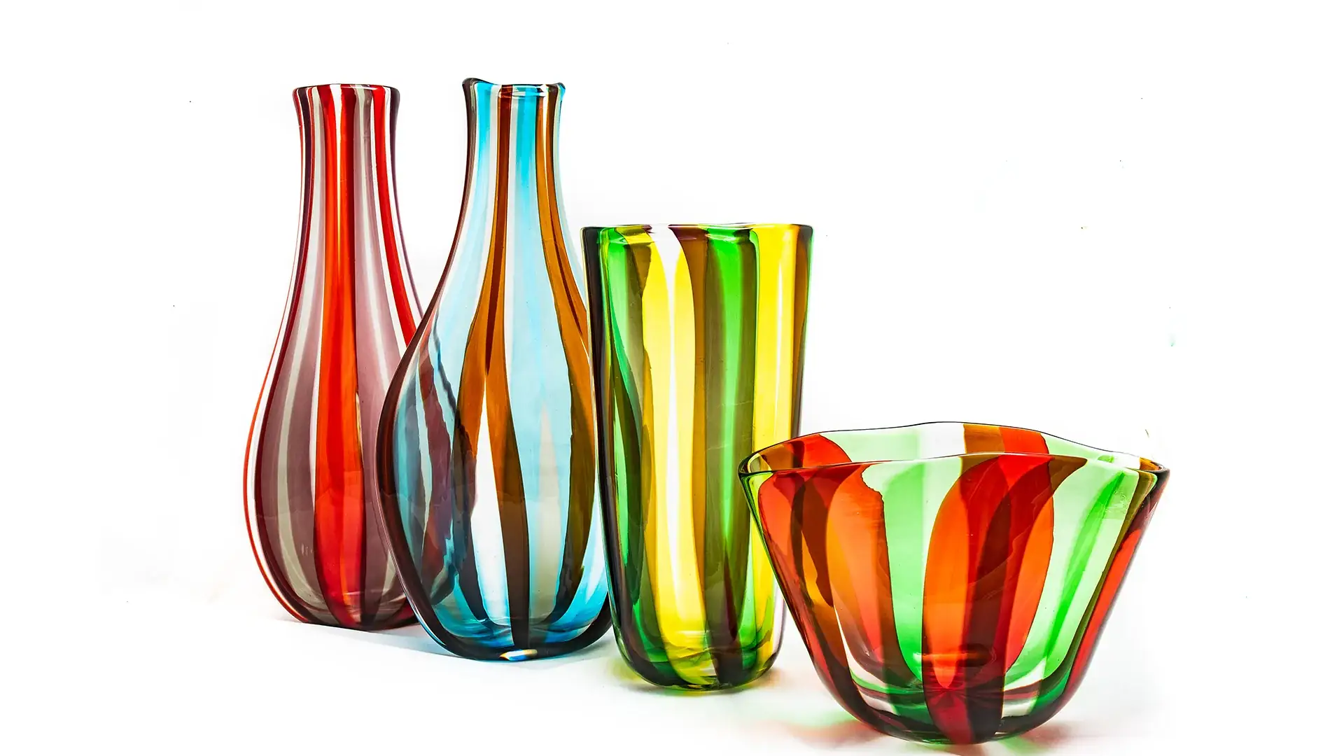Group of tall striped glass vases in red, blue, yellow, and green standing next to a matching striped glass bowl on a white background