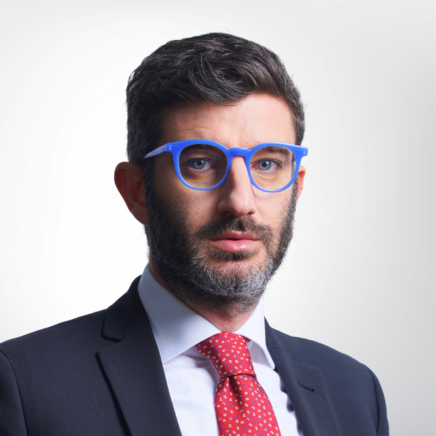 Professional portrait of a man with dark hair and beard wearing blue glasses, a navy suit, white shirt, and red tie against a light background.