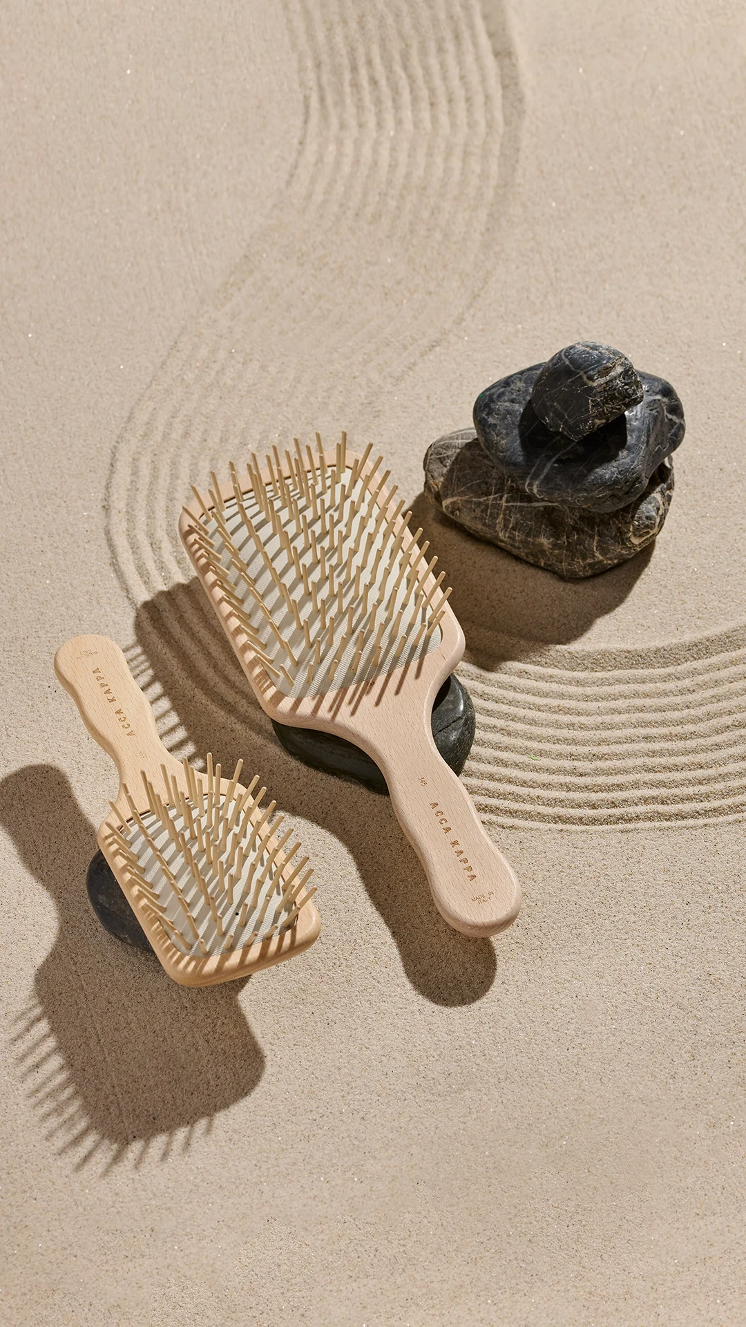 Two light wooden hair brushes resting on sand beside a small stack of smooth dark rocks, with a circular sand-rake pattern in the background.